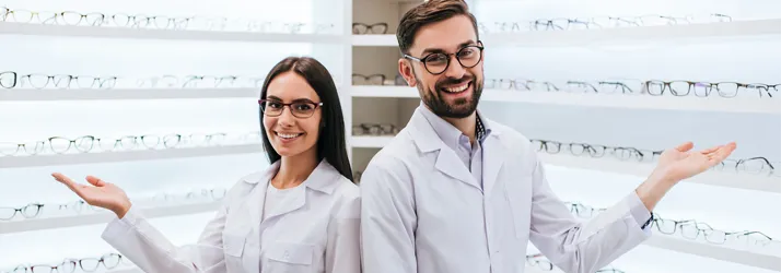 Two optometrists standing inside their office, gesturing to the many frames they have on display.